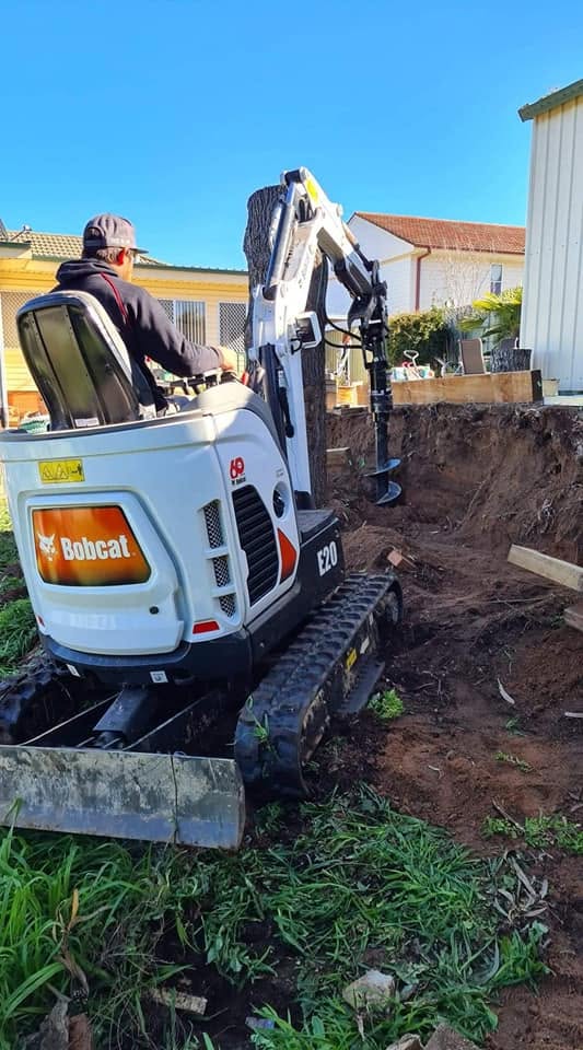 Markwicks Services crew member operating a Bobcat with auger attachment beside a tree